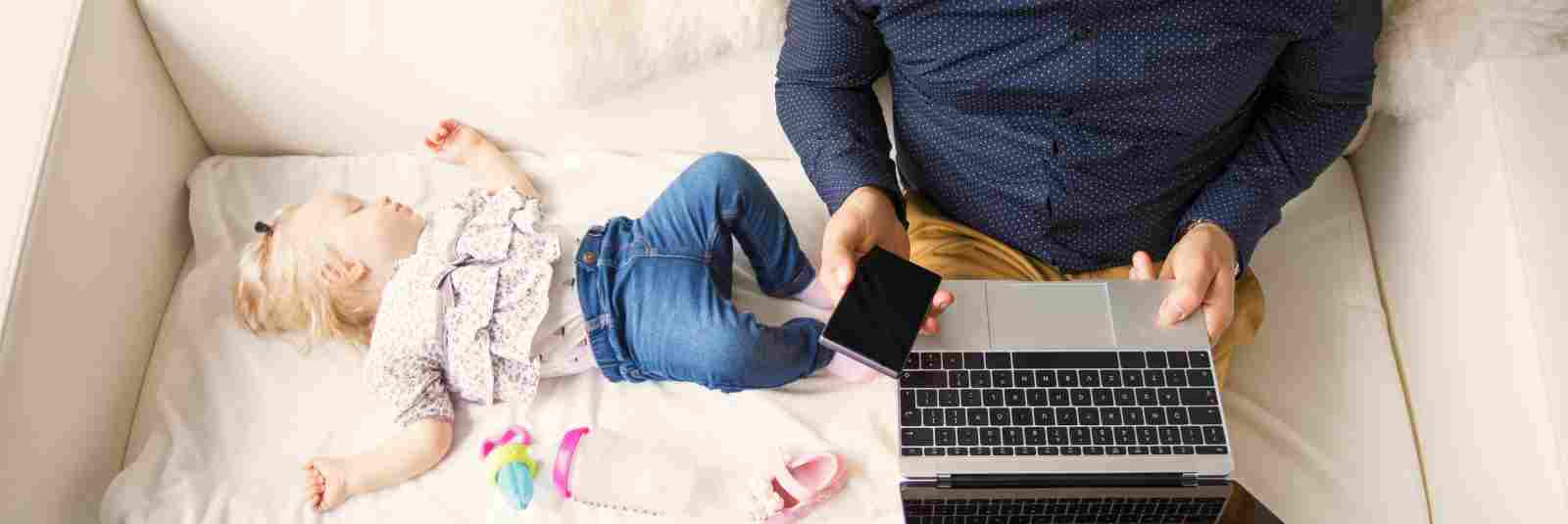 dad on laptop holding phone on couch with sleeping daughter